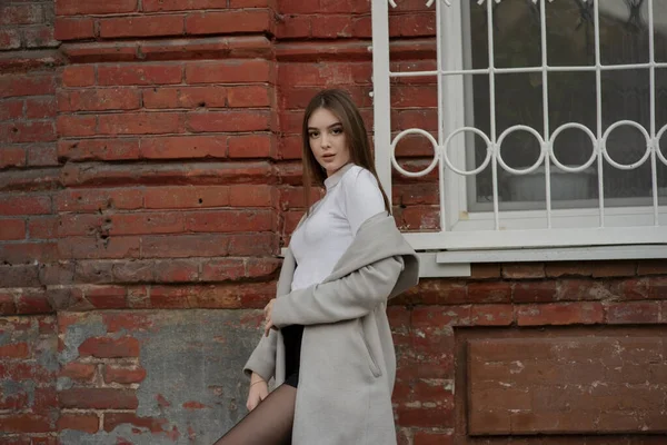 Jeune fille en manteau gris se dresse sur le fond d'une rue de la ville. Fille dans un manteau gris sur un fond gris . — Photo