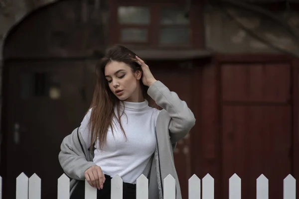 Niña con un abrigo gris se encuentra en el fondo de una calle de la ciudad. Chica en un abrigo gris sobre un fondo gris . —  Fotos de Stock
