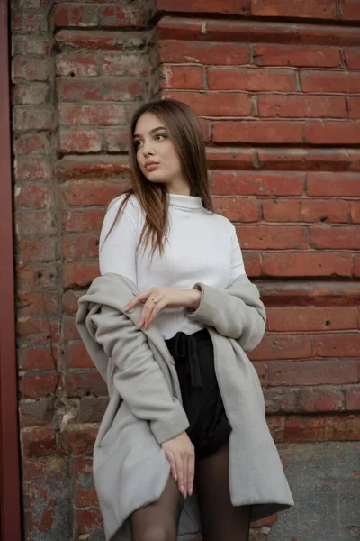Молодая девушка в сером пальто стоит на фоне городской улицы. Девушка в сером пальто на сером фоне . — стоковое фото