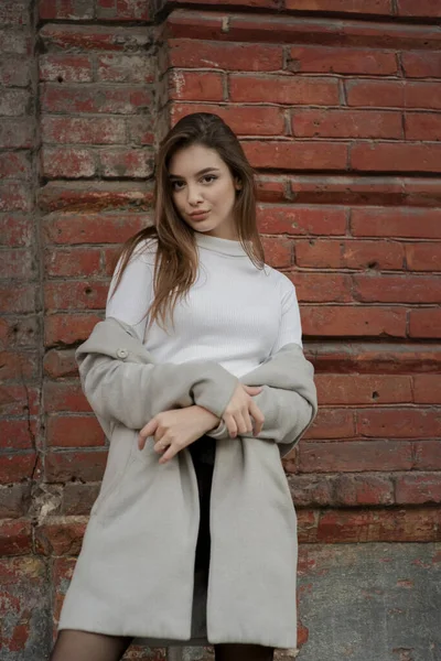 Giovane ragazza in un cappotto grigio si trova sullo sfondo di una strada della città. Ragazza in un cappotto grigio su uno sfondo grigio . — Foto Stock