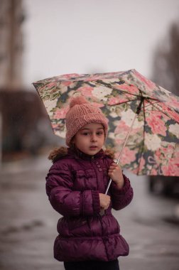Dışarıda şemsiyenin altında küçük bir kız, yağmurlu bir gün. Küçük kız yağmurun altında şemsiyeyle yürür. Yağmurlu bir günde pembe şapkalı ve mor ceketli bir çocuk.