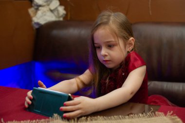 Bordo elbiseli küçük kız telefona bakıyor. 4-5 yaşlarında bir kız çocuğu kafede ya da evde akıllı telefondan ilginç bir şey izliyor. Bir çocuk telefonda haberleri izliyor..