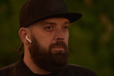 Yeşil arka planda sakallı bir adam. Siyah beyzbol şapkalı, siyah sakallı bir adam. Adam gün batımında vuruluyor. Kulağında kablosuz kulaklık olan bir adam. Tam yüz portresi.