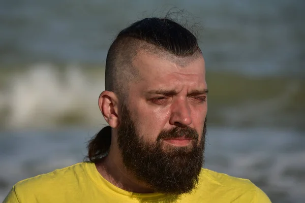 A young man wearing a yellow T-shirt and shorts walking along the sandy beach. Man in a yellow T-shirt with a beard on the background of the sea. Man and sea. Sea vacation summer concept.