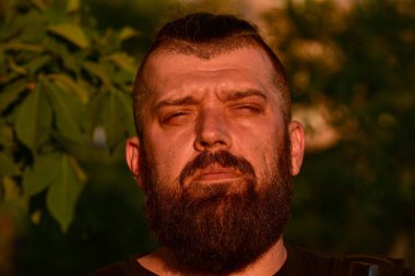 Yeşil arka planda sakallı bir adam. Gün batımında 40 yıllık sakallı bir adamın büyük yüz portresi. Tam yüz portresi. Siyah tişörtlü adam.
