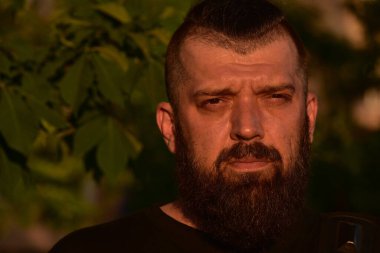 Yeşil arka planda sakallı bir adam. Gün batımında 40 yıllık sakallı bir adamın büyük yüz portresi. Tam yüz portresi. Siyah tişörtlü adam.