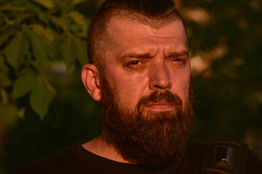 Yeşil arka planda sakallı bir adam. Gün batımında 40 yıllık sakallı bir adamın büyük yüz portresi. Tam yüz portresi. Siyah tişörtlü adam.