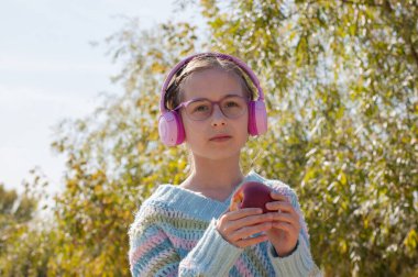 Parkta sırt çantalı, kulaklıklı kız öğrenci. Pembe kulaklıklı bir kız öğrenci okuldan sonra müzik dinler. Sonbahar okulu çocukluğu. 9 yaşında bir kızın portresi.