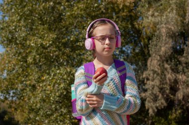 Parkta sırt çantalı, kulaklıklı kız öğrenci. Pembe kulaklıklı bir kız öğrenci okuldan sonra müzik dinler. Sonbahar okulu çocukluğu. 9 yaşında bir kızın portresi.