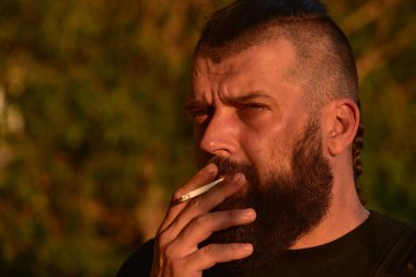 Yeşil arka planda sakallı bir adam. Gün batımında 40 yıllık sakallı bir adamın büyük yüz portresi. Tam yüz portresi. Siyah tişörtlü adam. Bir adam sigara içiyor..