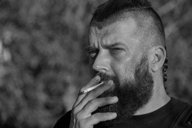Yeşil arka planda sakallı bir adam. Gün batımında 40 yıllık sakallı bir adamın büyük yüz portresi. Tam yüz portresi. Siyah tişörtlü adam. Bir adam sigara içiyor. Siyah beyaz fotoğraf