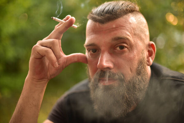 Человек с бородой на фоне зелени. Большой портрет бородатого мужчины 40 лет. Портрет полного лица. Человек в черной футболке. Мужчина курит сигарету
.