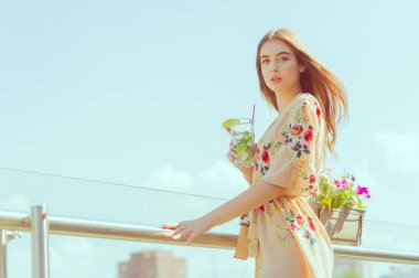 Güzel bir kız sıcak ve bunaltıcı bir yaz gününde şehrin arka planında buzlu mojito içer. Açık terasta bir kafede seksi bir esmer. Kız hayattan zevk alıyor. Çiçekli kıyafetler.