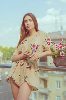 Güzel bir kız sıcak ve bunaltıcı bir yaz gününde şehrin arka planında buzlu mojito içer. Açık terasta bir kafede seksi bir esmer. Kız hayattan zevk alıyor. Çiçekli kıyafetler.