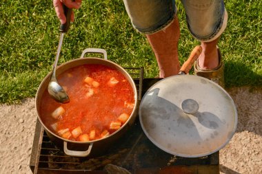 Avludaki kazıkta çorbayla birlikte dökme demir kazan. Yaz günü ve çiçek tarhının yanındaki temiz havada akşam yemeği. Çorba kazıkta bir kazanda hazırlanır. Kazıkta çorba pişirmek.