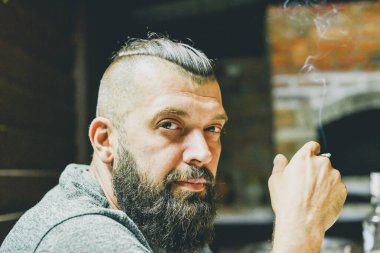 Siyah saçlı ve sakallı yeşil tişörtlü bir adam. Esmer adam. Şöminenin arkasında sakallı bir adam var. Örgülü ve yandan traş olmuş bir adam için şık bir saç stili.