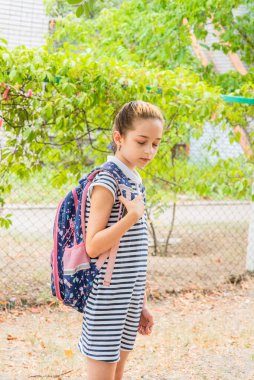 Okula yürümeye hazırlanan küçük bir kız. Okul üniformalı, sırt çantalı bir kız. Sonbaharda okul bahçesinde genç.