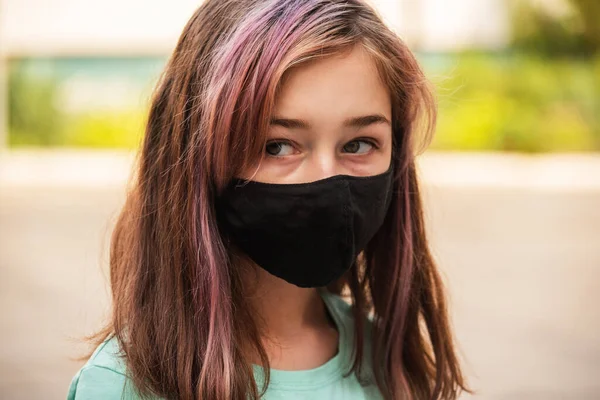 A young girl wears a face mask that protects against the spread of coronavirus disease. Close- up of teenager in an antiviral mask. Girl in a protective mask outdoors. Pandemic, virus, coronavirus