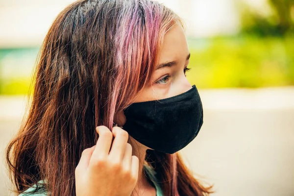 A young girl wears a face mask that protects against the spread of coronavirus disease. Close- up of teenager in an antiviral mask. Girl in a protective mask outdoors. Pandemic, virus, coronavirus