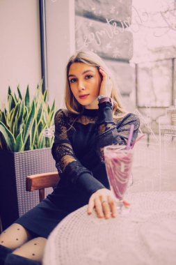 Meyveli buzlu güzel genç bayan. Kafede smoothie içen bir kız. Siyah elbiseli kız. Doğal saçlı bir kız. Kız ve kafede rahatlama, sağlıklı yiyecekler, vitaminler. Sakin kavram