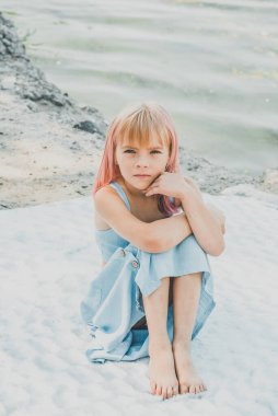 Nehrin kenarında oturan sevimli küçük kız. Nehir kenarındaki küçük kız, ekoseli bir elbiseyle. Çocukluk kavramı