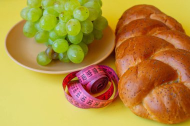Şerit mezura ve sarı arka planda haşhaş tohumlu çörek. Sağlıklı gıda, diyet, kilo kaybı ve zayıflık kavramı. Tabakta üzümler