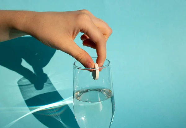 Mavi arka planda, bir el eriyen bir aspirin tabletini bir bardak suya düşürür..