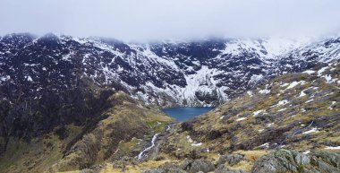 Kışın Snowdon'da yapılan bir yürüyüş sırasında yakalanan göl, engebeli vadi ve karamsar gökyüzünü n üstünden çarpıcı dağ manzarası (Snowdonia Ulusal Parkı, Galler, Birleşik Krallık)