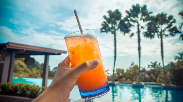 Close up of an orange tropical cocktail in hand by the pool in summer, with blurred palm trees and turquoise blue sky in the background. (Summer, travel and vacation concept)