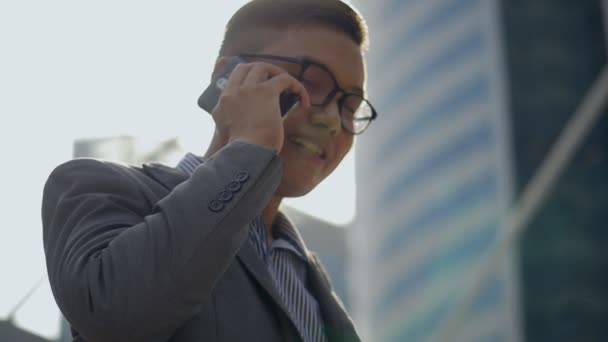 肖像画ハンサムなアジアのビジネスマンは 都市部で友人と電話を話します 眼鏡をかけたアジア人の男性は 大きなオフィスビルの近くの通りで電話を使います スローモーション撮影 — ストック動画