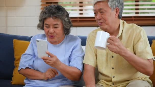 スマートフォンを使ってビデオチャットをしているアジアのシニアカップル 自宅のソファに座ってソーシャルメディアのコミュニケーションを共有します スローモーション — ストック動画