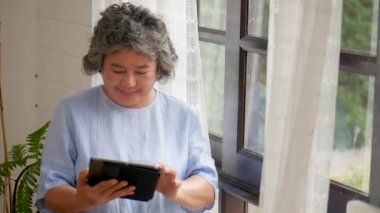 Portre asya kıdemli kadın tablet online alışveriş arama kullanarak. Evde pencere nin yanında sosyal medya iletişimini paylaşma