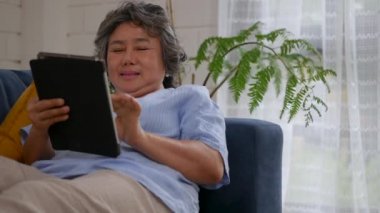 Portre asya kıdemli kadın tablet online alışveriş arama kullanarak. Evde kanepede yatarken sosyal medya iletişimini paylaşmak.
