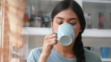 Genç Asyalı kadın mutfakta pencerenin yanında tablet kullanıyor ve kahve içiyor..