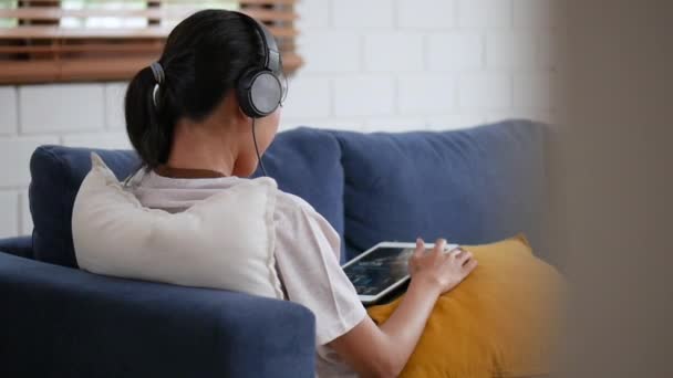 リビングルームで音楽を聴くヘッドフォンでソファの上に座っている美しい若いアジアの女性を肖像 — ストック動画