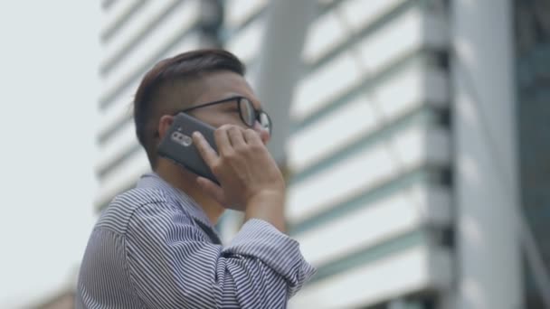 スローモーションの肖像画アジアのビジネスマンは 電話に答えます 眼鏡をかけたアジア人の男性が大きなオフィスビルの近くの通りで電話を使う — ストック動画