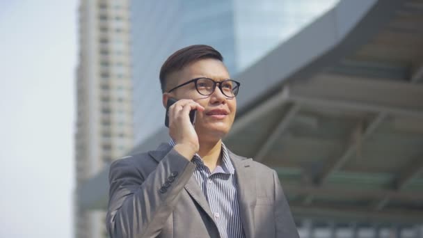 都会を歩く友人と電話をかけるハンサムなアジアのビジネスマンの肖像画 眼鏡をかけたアジア人の男性は 大きなオフィスビルの近くの通りで電話を使います スローモーション撮影 — ストック動画