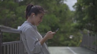 Gülümseyen portre güzel genç Asyalı kadın fitness koşucusu bir kamu parkta müzik dinlerken cep telefonu kullanarak ayakta.