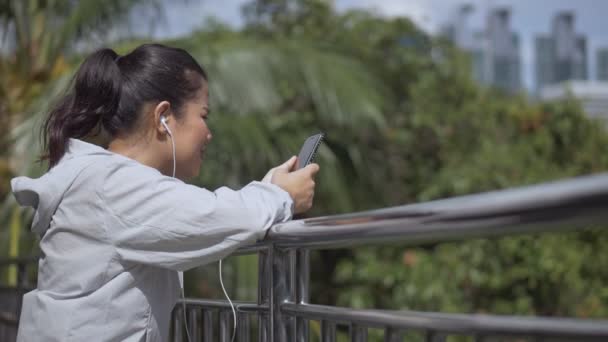 微笑的肖像美丽的年轻亚洲妇女健身亚军站在使用手机听音乐在公共公园 — 图库视频影像