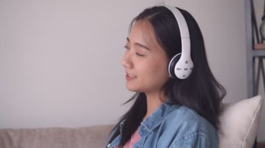 Evde oturma odasında kanepede otururken kulaklık müzik dinlerken çekici genç Asyalı kadın.