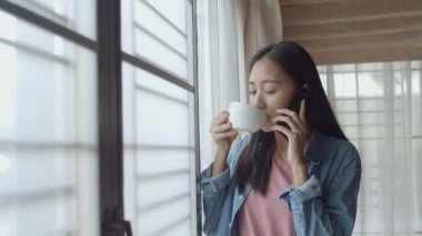 Çekici portre gülümseyen genç Asyalı kadın kahve içmek ve ev ofisinde pencerenin yanında duran arkadaşları ile Telefonda konuşurken.