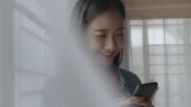 肖像微笑的年轻亚洲女商人拿着打字手机 在智能手机的社交媒体上滚动浏览社交媒体源 站在家庭办公室的窗户旁 — 图库视频影像