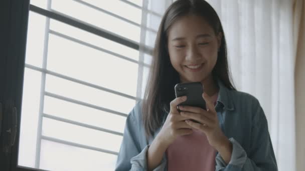 携帯電話をタイピングし ホームオフィスの窓の横に立っているスマートフォンでソーシャルメディアフィードをスクロールしてスクロールを持つ若いアジアのビジネスウーマンの肖像画 — ストック動画