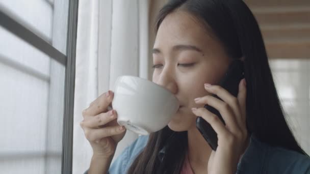 特写迷人的肖像微笑年轻的亚洲妇女喝咖啡 并与站在家庭办公室窗口旁边的朋友电话交谈 — 图库视频影像