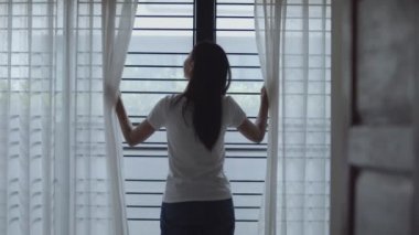 Çekici genç Asyalı kadın evde odada perdeleri açma pencereden dışarı bakıyor ve sabah tatil saatinde germe.