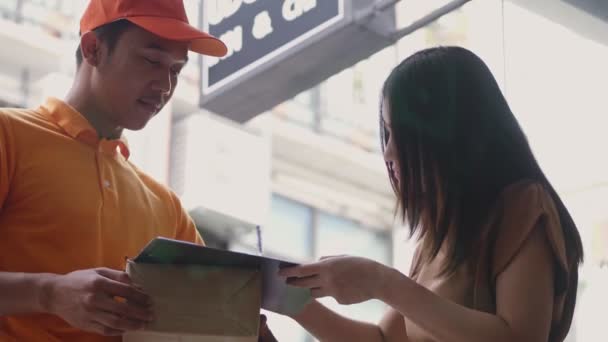 アジアの若い女性がクリップボードに署名を入れている間 段ボール箱を持つ笑顔の若い配達人 — ストック動画