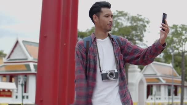 タイのストリート巨大スイングで友人とスマートフォンのビデオチャットを使用して立ってハンサムな若いアジアの男のソロ観光客の肖像画 アジアの男性ブロガー幸せなレジャーライフスタイル旅行を楽しむ — ストック動画
