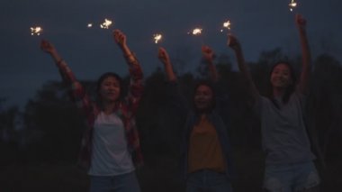 Bir grup genç Asyalı kadın ellerinde maytap tutarak yeni yılı kutluyorlar doğada kamp yapıyorlar birlikte bira içerek eğleniyorlar..