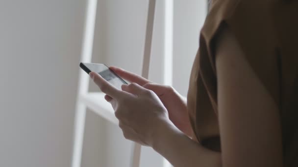 Nahaufnahme junge asiatische Geschäftsfrau hält Handy in der Hand und blättert durch Social-Media-Feed in Smartphone neben einem Fenster im Homeoffice stehen.