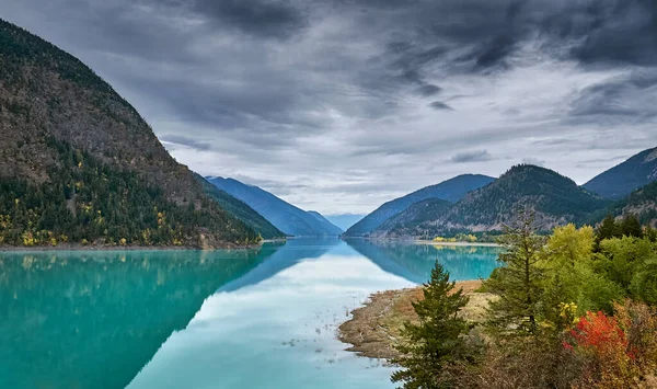 Turquoise mountain Carpenter lake in autumn. Mountains with coniferous forest around the lake.  Red and yellow autumn trees in the foreground. Autumn forest landscape. BC, Canada